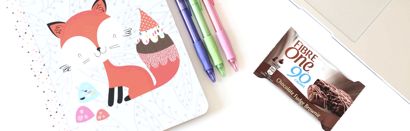 A table top with a notebook, a selection of pens and a Fibre One 90 Calorie chocolate fudge brownie.